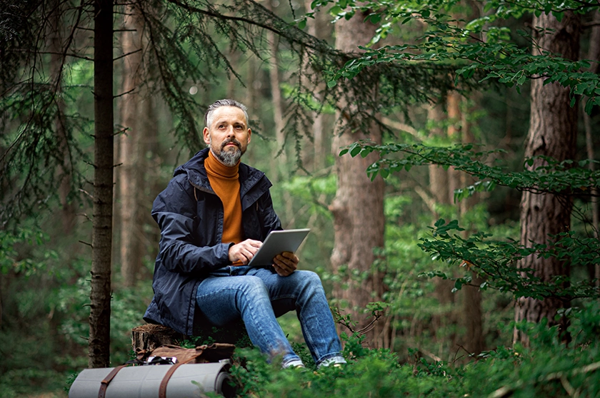 A man sitting outdoor in teh forest working on his iPad. This picture illustrates how outdoor office work can look like.